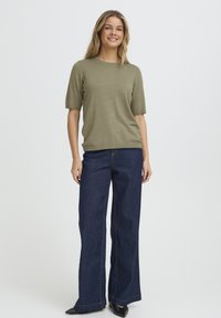 Olivzaļš īsroku džemperis, kombinēts ar tumši ziliem plaša piegriezuma džinsiem. Apģērbam ir vienkāršas līnijas un brīvs siluets, melnas plakanas kurpes.