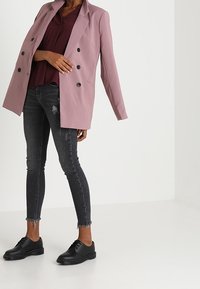 Blazer oversize di colore rosa chiaro con due bottoni neri, abbinato a un top bordeaux e jeans skinny neri strappati, completato da scarpe nere con lacci.