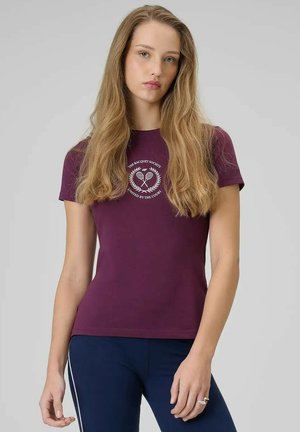 Młoda kobieta z długimi włosami, ubrana w fioletowy t-shirt z logo rakiety tenisowej i granatowe spodnie na jednolitym tle.