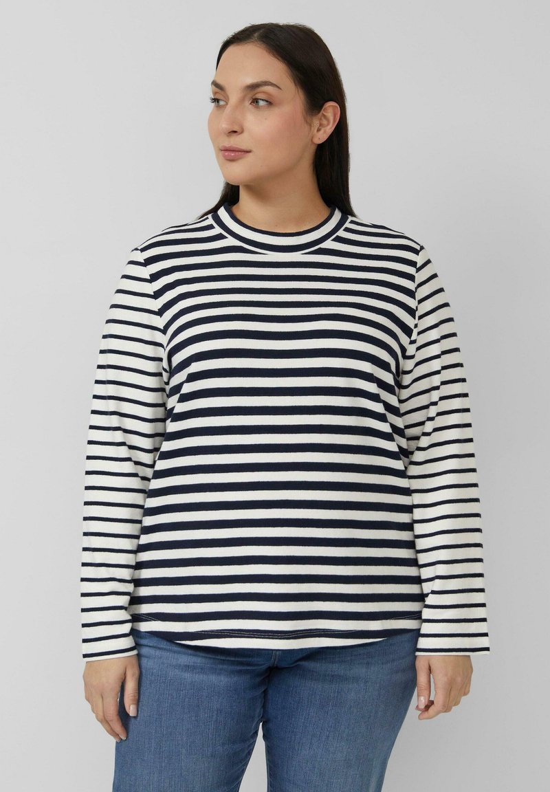 Frau trägt ein langärmeliges Shirt mit horizontalen marineblauen und weißen Streifen und blaue Jeans, schaut leicht nach links vor einem schlichten Hintergrund.