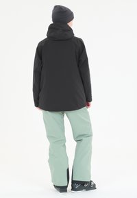 Mustat vedenpitävät takki hupulla, yhdistettynä vaaleanvihreisiin housuihin ja mustiin hiihtokenkään. Sujuva pinta ja toiminnallinen muotoilu.