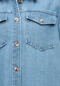 Chemise en denim bleu clair à l'avant avec deux poches poitrine boutonnées et quatre boutons beiges visibles au centre.