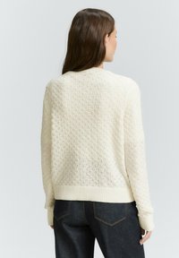 Maglione bianco con texture, orlo e polsini a coste, caratterizzato da un motivo tessuto. Il modello indossa jeans in denim scuri.