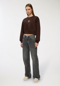 Felpa corta marrone con logo frontale, abbinata a jeans grigi sbiaditi a vita alta efit morbido, e sneakers beige. Design semplice, senza fantasie.