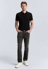 Juodas polo marškinėlis su raudonos spalvos apykaklės akcentais, derinamas su tamsiai pilkais džinsais ir baltais sportbačiais. Modelis stovi su rankomis kišenėse, priešais pilką foną.