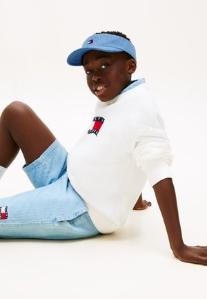 Garçon assis par terre portant un pull blanc avec un logo, un short en jean bleu clair et une casquette visière bleue assortie, regardant vers la caméra.
