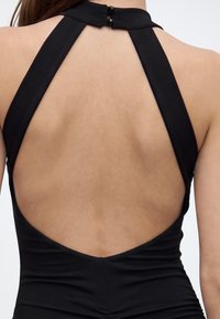 Femme portant une robe noire dos-nu avec deux boutons au niveau du cou sur un fond uni.