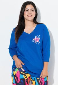 Blaues V-Ausschnitt Langarmshirt mit einem rosa Stern-Graphic und weißen Akzenten, kombiniert mit bunten Leggings im abstrakten Muster.
