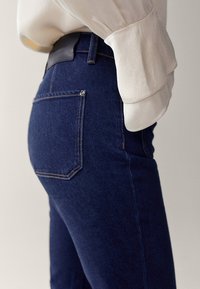 Mörkblå denimjeans med hög midja, med två bakfickor och kontrasterande sömmar. Bärs tillsammans med en ljus, strukturerad blus.