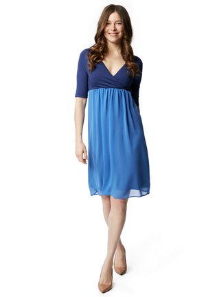 Vestito blu con scollatura a V, maniche corte e vita impero. La gonna è realizzata in un tessuto leggero e fluido. Indossato con tacchi nudi.