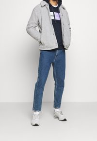 Grå fleecetröja med tryckknappar, lager-på-lager över en marinblå hoodie med flerfärgade ränder. Blå jeans i denim och vita sneakers.