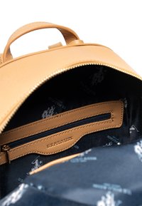 Intérieur d'un sac à dos en cuir tan avec une poche zippée. Doublé d'un tissu sombre présentant des logos blancs répétitifs de U.S. Polo Assn.