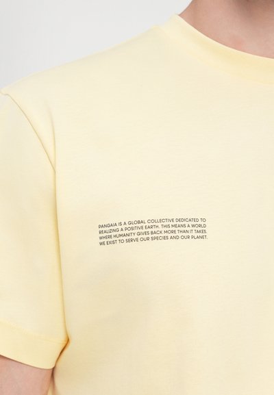 Tricou din bumbac galben deschis, cu guler rotund. Prezintă text imprimat în întuneric pe partea stângă a pieptului, punând accent pe sustenabilitate.
