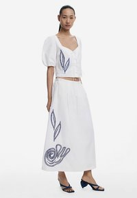 Ținută albă în două piese, formată dintr-un top scurt cu mâneci bufante și modele brodate cu frunze albastre, asortată cu o fustă lungă cu broderii similare.