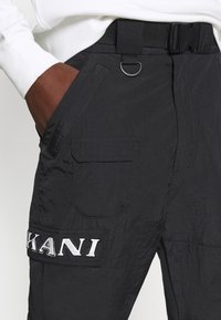 Gros plan sur un pantalon cargo noir avec poche plaquée, lettrage du logo en argent, une ceinture noire avec boucle en plastique et une main dans la poche avant.