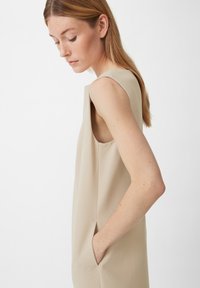 Beiges ärmelloses Kleid mit strukturiertem Finish, seitlichen Taschen und gerader Silhouette. Der Stoff wirkt leicht und atmungsaktiv.