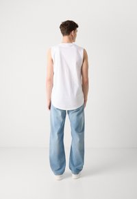 Camiseta sin mangas blanca con dobladillo redondeado, combinada con vaqueros de pierna ancha azul claro y zapatillas blancas, vista desde la parte posterior.