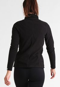 Vrouw in een zwarte fleece jack met lange mouwen en zwarte leggings, staande met haar rug naar de camera tegen een neutrale achtergrond.