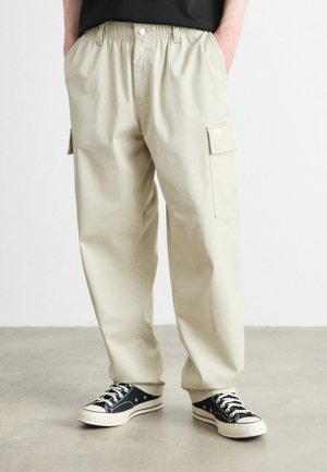 Beige losse cargobroek met zijzakken, gedragen met zwarte laag uitgesneden canvas sneakers en een zwart shirt, staand op een betonnen vloer.