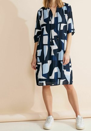 Kobieta w sukience do kolan z abstrakcyjnym wzorem w kolorach granatowym, białym i jasnoniebieskim, w połączeniu z białymi trampkami, stojąca na neutralnym tle.