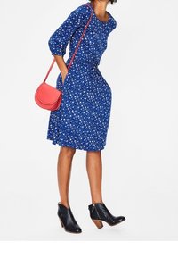 Blå blomsterklänning med trekvartsärmar, som har en figurnära midja och en svängande kjol. I kombination med en röd axelväska och svarta ankelstövlar.