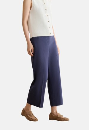 Person trägt ein ärmelloses weißes Strick-Top mit Knöpfen, weite marineblaue kurze Hosen und braune Wildlederloafers mit goldenen Details.