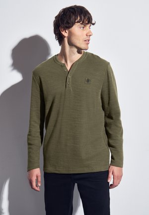 Camisa henley de manga larga en verde oliva con un acabado texturizado, que cuenta con una tapeta de tres botones y un pequeño parche con el logo en el pecho.