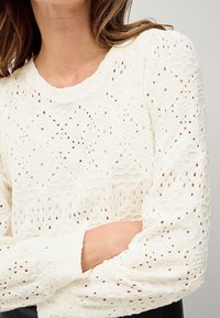 Top en dentelle blanche avec un motif en diamant texturé, des manches longues et un col rond, comportant des découpes visibles sur tout le tissu.
