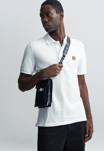 Chemise polo blanche avec une petite broderie de renard orange sur la poitrine, accompagnée d'un sac en cuir noir avec un fermoir argenté et une bandoulière ajustable.