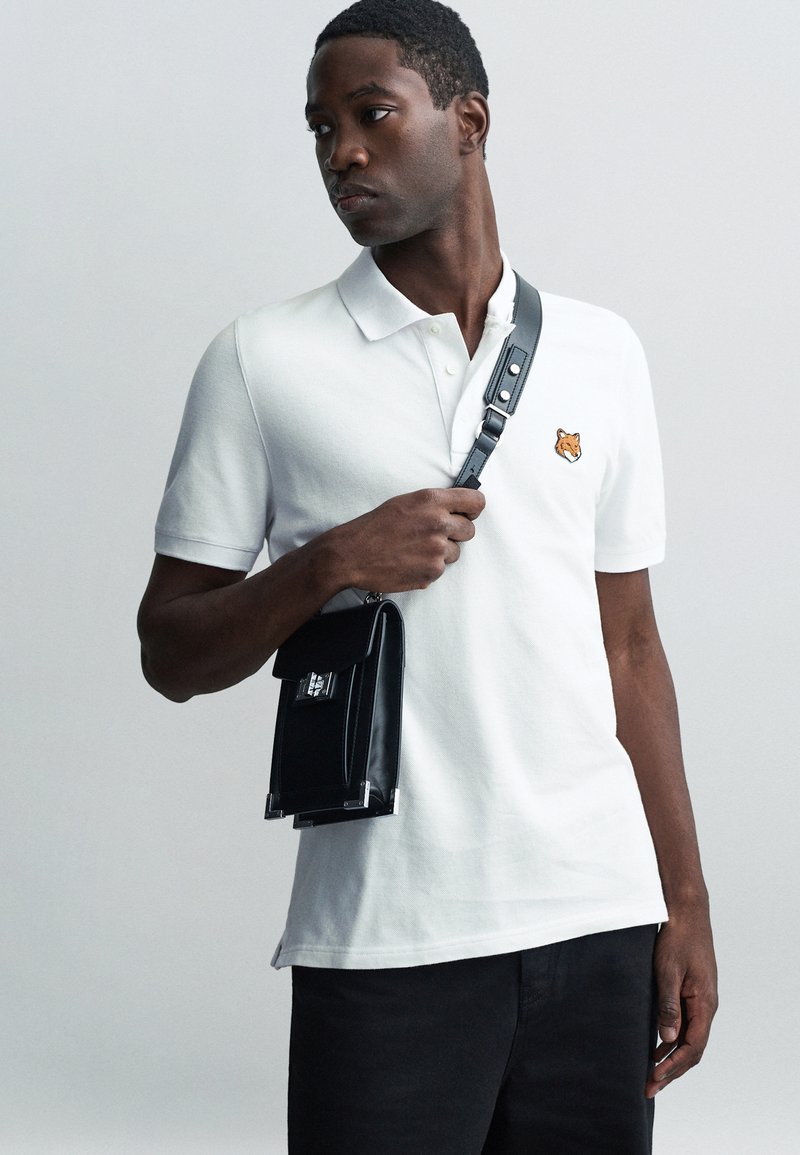 Tricou polo alb cu un mic brodat de vulpe portocalie pe piept, asortat cu o geantă din piele neagră cu un clasp argintiu și curea ajustabilă.