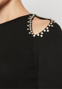 Svart långärmad topp med ribbad textur och skärning vid axlarna, prydd med kristall-liknande dekorationer längs halsringningen.