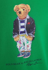 Camiseta verde con un gráfico de un oso con un blazer negro, camisa a rayas, jeans decorados y zapatos azules, con un reloj en su muñeca.