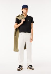 Sort V-hals t-shirt, hvide lige ben jeans, olivengrøn jakke over skulderen, marineblå hat og mønstrede sneakers.