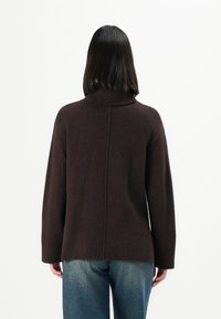 Personne avec des cheveux noirs raides portant un pull à col roulé marron et un jean bleu, de dos contre un fond blanc uni.