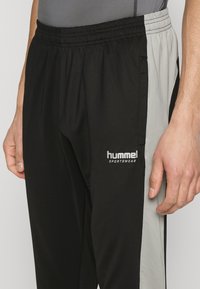 Schwarze und graue Sporthose mit elastischem Bund, ausgestattet mit einer glatten Textur und einem bedruckten Logo am Oberschenkel.