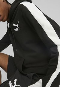Felpa nera con strisce bianche sulle maniche, dotata di tasca frontale e logo Puma bianco. Tessuto morbido con finitura testurizzata.