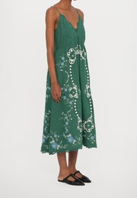 Zelené midi šaty s hlubokým výstřihem do V, vyšívanými květinovými vzory, krajkovým lemem a tenkými ramínky, doplněné o černé šlapky.