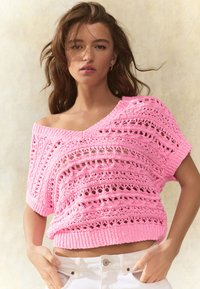 Pink cropped knit sweater with openwork design, featuring short sleeves and a wide neckline, paired with white trousers.