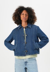 Jeans jakna v temno modri barvi z ovratnikom, sprednjim zapiranjem na gumb, dvema stranskima žepoma in sproščenim krojem, nosena čez svetlo zeleno majico.