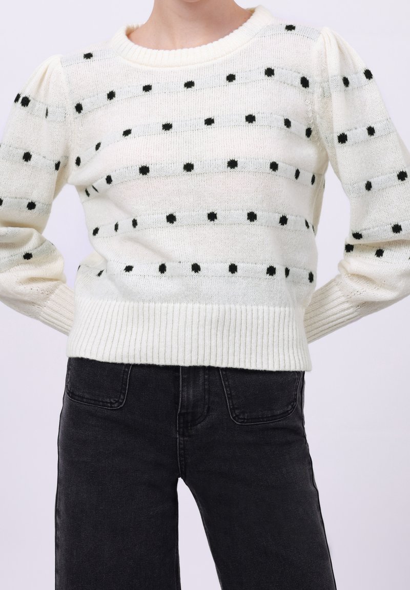 Pull en maille blanche avec des rayures grises horizontales et des pois noirs espacés régulièrement, porté avec un jean noir taille haute, mains sur les hanches.