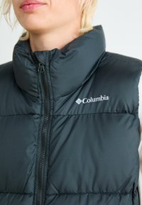 Crna, pufasta prsluk s visokim ovratnikom i prednjim rajsferšlusom. Sadrži Columbia logotip na lijevoj strani prsnog koša. Napravljen je od laganog materijala.