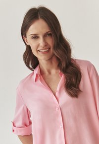 Lichtroze blouse met een knoopsluiting, voorzien van een kraag en opgerolde mouwen. Gemaakt van een gladde, lichte stof.