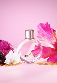 Clear round glass perfume bottle with a silver cap, surrounded by pink hibiscus flowers, blackberries, and amber resin on a pink background.