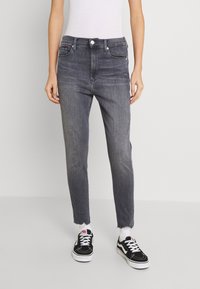 Persona vistiendo unos pantalones skinny de tiro alto en negro deslavado con dobladillo sin rematar, calcetas blancas, zapatillas en blanco y negro, y una blusa sin mangas blanca.