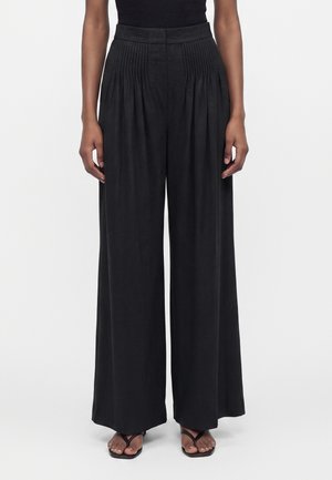 Personne portant un pantalon large noir taille haute avec plis à la taille et des sandales noires à lanières sur fond blanc.
