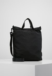 Borsa tote rettangolare nera con manici corti e tracolla regolabile, posizionata su una superficie bianca contro uno sfondo grigio.