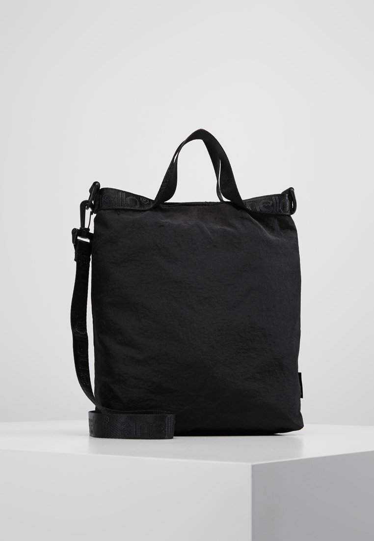 Borsa tote rettangolare nera con manici corti e tracolla regolabile, posizionata su una superficie bianca contro uno sfondo grigio.