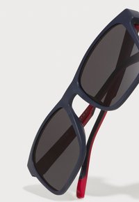 Ochelari de soare cu ramă din plastic mat de culoare navy, lentile întunecate și accente roșii pe brațe. Formă pătrată cu margini unghiulare.
