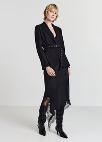 Veste noire ajustée avec ceinture et détails en boutons, superposée à une longue robe noire avec des accents en dentelle et des bottes noires à talons.