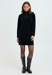 Zwarte corduroy jurk met hoge kraag en halfrits sluiting, voorzien van lange mouwen en een relaxte pasvorm, gecombineerd met kniehogere zwarte laarzen.